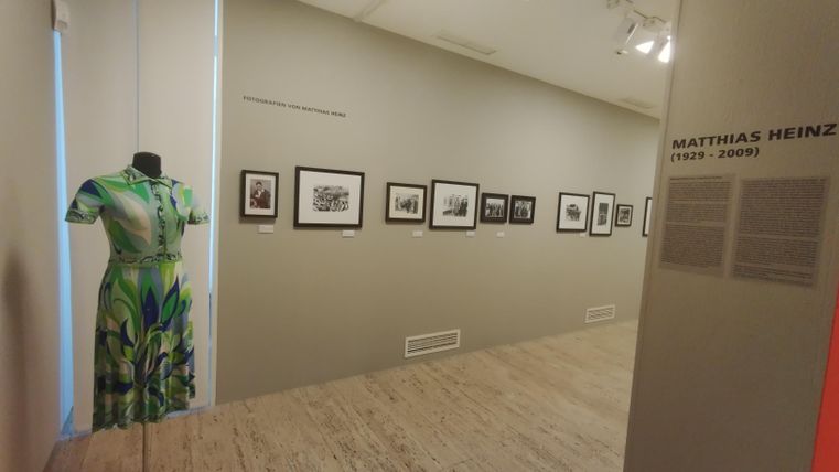 Ein Ausstellungsraum mit einem grünen Kleid auf einem Stand und mehreren Bildern an der Wand. Die Ausstellung zeigt Werke von Matthias Heinz.