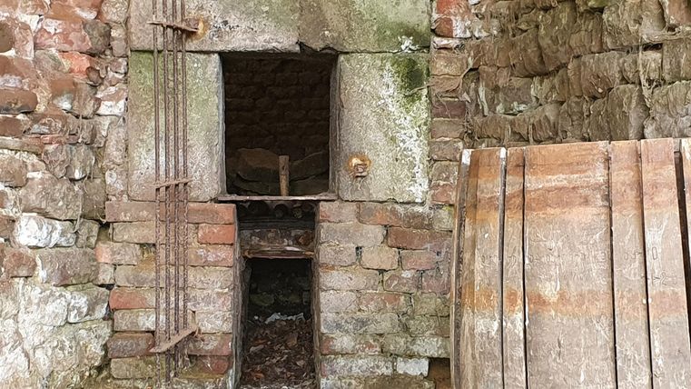 Eine alte, gemauerte Struktur mit geöffneten Toren. Daneben liegen einige Holzplanken auf dem Boden.