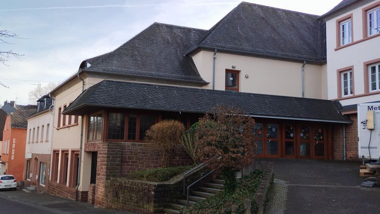 Ein modernes Gebäude an einer ruhigen Straße, mit einer schrägen Dachkonstruktion. Die Fassade ist aus Stein und Holz, umgeben von kleinen Bäumen und Büschen.