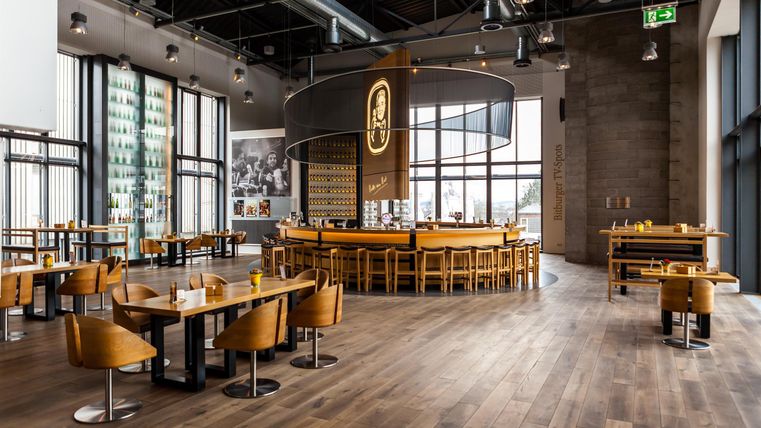 Ein modernes Restaurant mit einer zentralen Bar und Holzmöbeln. Große Fenster sorgen für viel Tageslicht und eine einladende Atmosphäre.