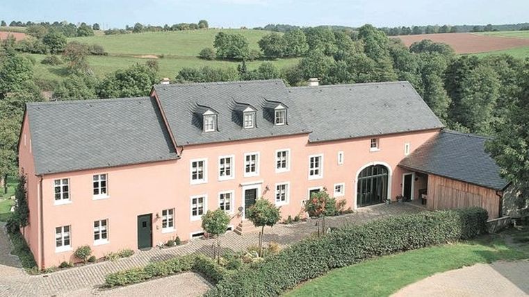 Ein schönes, rosa Haus mit einem großen Garten und sichtbaren Bäumen in der Umgebung. Die Landschaft ist grün und hügelig.