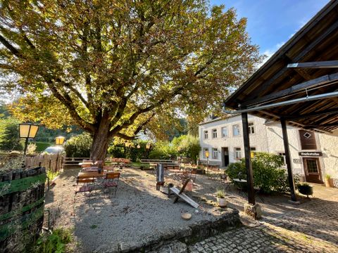 A cozy outdoor area with tables and chairs under a large tree. In the background, a building can be seen that creates an inviting atmosphere.
