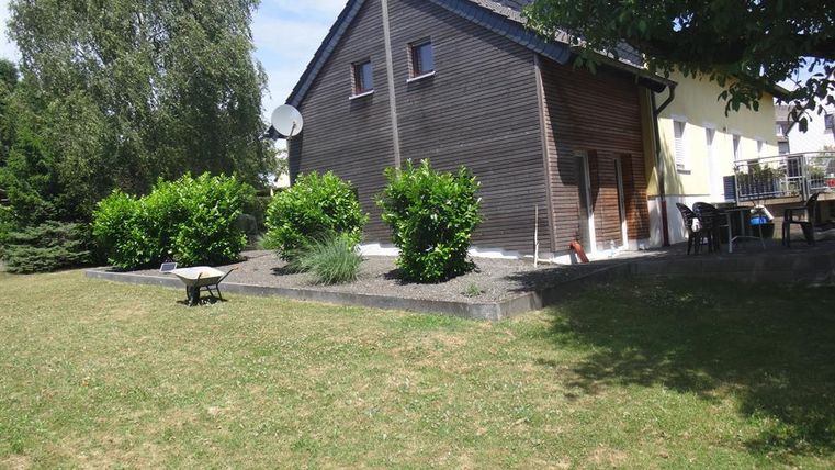 Une maison en bois avec un jardin soigné et quelques arbres. Le soleil brille et le ciel est dégagé.
