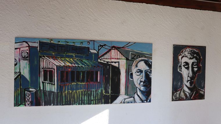 Zwei gemalte Bilder an einer weißen Wand. Das linke Bild zeigt ein Gebäude mit einem Gesicht, während das rechte Bild eine portraitartige Darstellung einer Person zeigt.