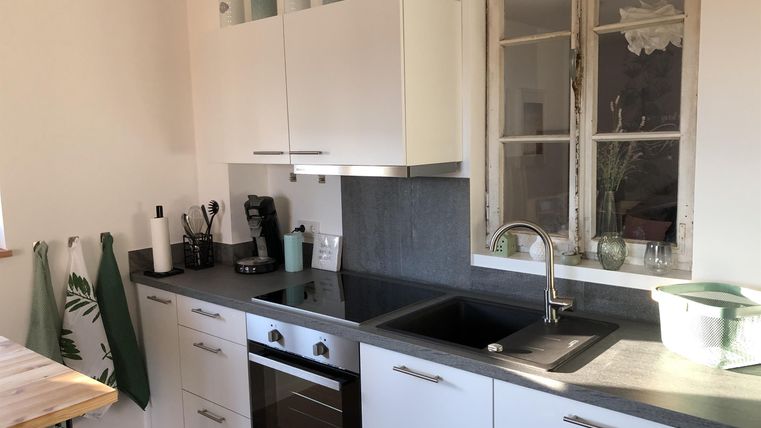 Eine moderne Küche mit weißen Schränken und schwarzer Arbeitsplatte. Im Hintergrund sind ein Fenster und einige dekorative Gegenstände zu sehen.