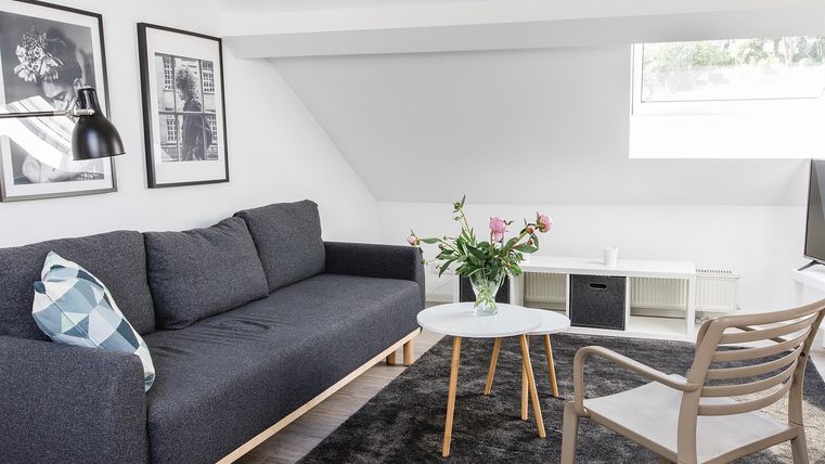 Ein stilvolles Wohnzimmer mit einem grauen Sofa und einem kleinen Tisch. An der Wand hängen Bilder, und ein Blumenstrauß steht auf dem Tisch.