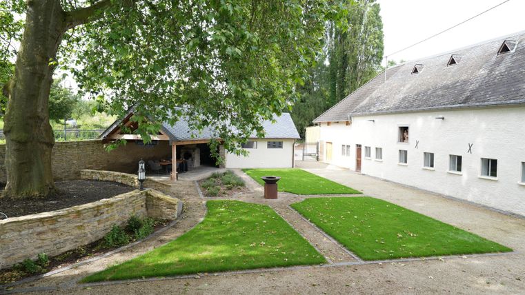 Eine ruhige Hofanlage mit grünem Rasen, Bäumen und einem Nebengebäude. Im Hintergrund sind alte Stallgebäude zu sehen.