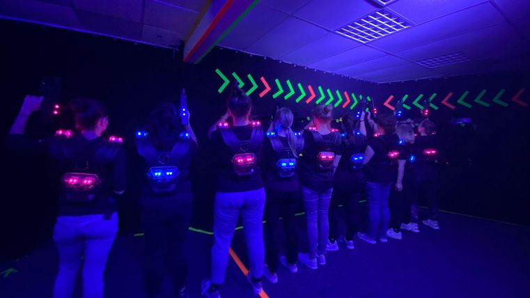 Un groupe de personnes se tient dans une pièce sombre avec des lasers lumineux. Ils portent des gilets spéciaux et se préparent à un jeu de laser.