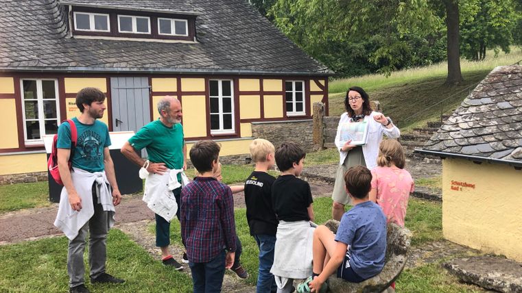 Eine Gruppe von Kindern hört einer Frau zu, die etwas erklärt. Im Hintergrund ist ein historisches Gebäude in einer grünen Umgebung zu sehen.
