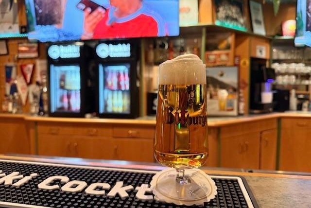 Een glas bier staat op de bar. Op de achtergrond zijn drankautomaten en televisies te zien.