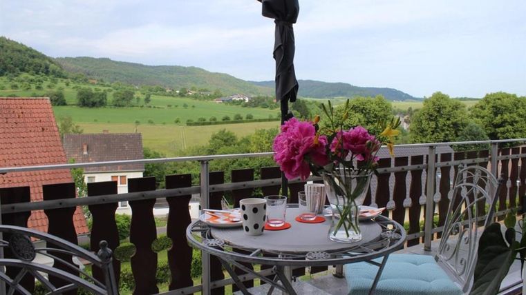 Ein gemütlicher Balkon mit einem Tisch und Stühlen, auf dem eine Vase mit bunten Blumen steht. Im Hintergrund sind grüne Hügel und eine malerische Landschaft zu sehen.