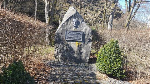 Ein Gedenkstein in einer ruhigen Umgebung, umgeben von Büschen und kleinen Bäumen. Auf dem Stein befindet sich ein Relief und eine Inschrift.
