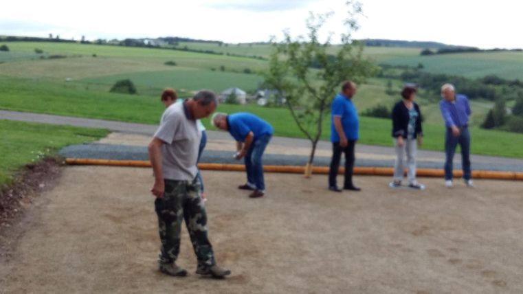 Eine Gruppe von Menschen spielt Boule auf einem schottrigen Platz im Freien. Im Hintergrund ist eine grüne Landschaft mit Hügeln zu sehen.