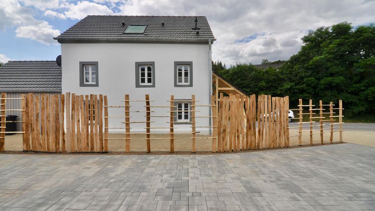 Ein modernes, einseitig weißes Haus mit einem grauen Dach. Es ist von einem Holzzaun umgeben, und im Hintergrund sind Bäume zu sehen.