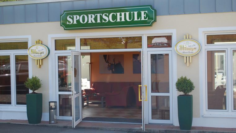 Een uitnodigende ingang van een sportschool met grote ramen. De gevel is licht en modern vormgegeven.