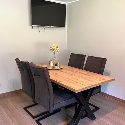 Ein moderner Essbereich mit einem Holztisch und vier dunklen Stühlen. An der Wand hängt ein Fernseher, und auf dem Tisch steht eine dekorative Vase.