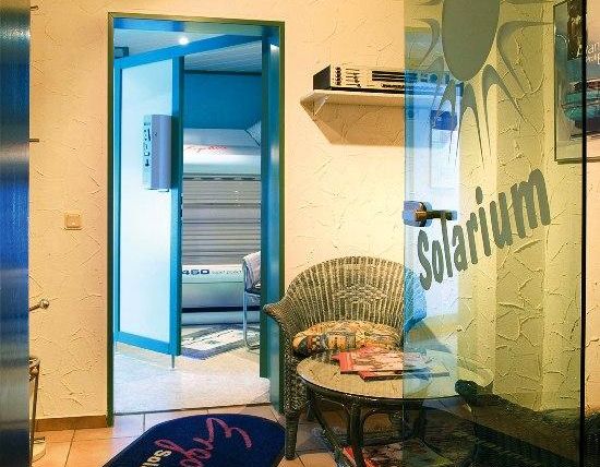 Ein Empfangsbereich eines Solariums mit einem gemütlichen Stuhl und einem Tisch. Hinter einer Glastür ist das Solariengerät sichtbar.