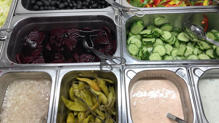 Eine Auswahl an frischem Gemüse und Beilagen, darunter Oliven, Gurken und Paprika. Es gibt auch verschiedene Saucen und Dips in separaten Behältern.