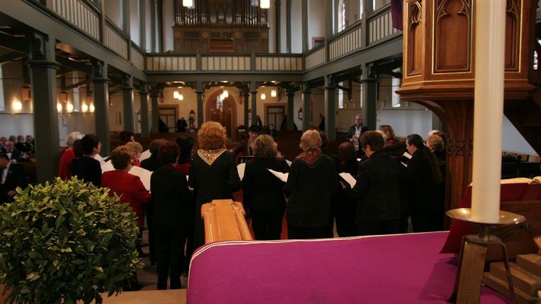 Eine Gruppe von Sängern steht in einer Kirche und singt. Im Vordergrund ist ein violettes Tuch und eine kleine Pflanze zu sehen.