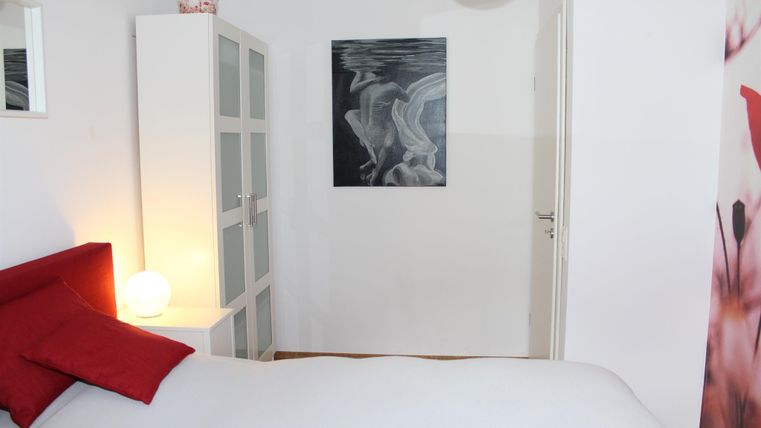 Ein gemütliches Schlafzimmer mit einem roten Bett und dekorativen Kissen. An der Wand hängt ein modernes Gemälde, und der Raum ist hell und einladend.