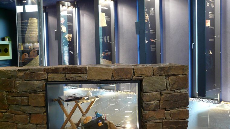 Ein Ausstellungsraum mit mehreren Vitrinen, die historische Objekte zeigen. Im Vordergrund ist ein Steinsockel mit einem Tisch und verschiedenen Sammlerstücken sichtbar.