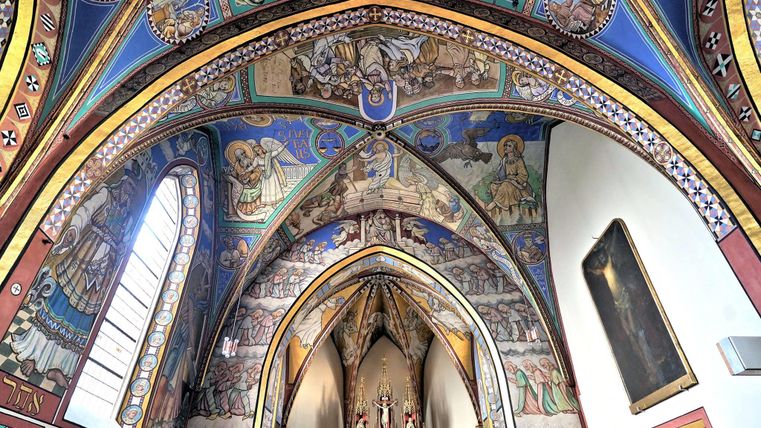 Eine kunstvoll verzierte Decke mit bunten Wandmalereien in einer Kirche. Licht strömt durch die Fenster und betont die Details der Architektur.