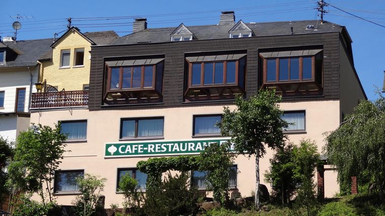 Ein Café-Restaurant in einem zweigeschossigen Gebäude mit großen Fenstern. Grünpflanzen und Bäume umgeben das Haus, das von einem klaren Himmel profitiert.
