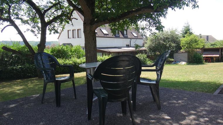 Un coin salon confortable dans le jardin avec des meubles en plastique noirs sous un arbre. À l'arrière-plan, on peut voir des plantes vertes et un bâtiment.