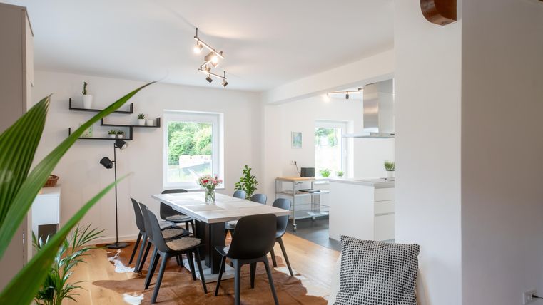 Un salon moderne avec une table à manger et des chaises noires. De grandes fenêtres laissent entrer beaucoup de lumière du jour et des plantes créent une atmosphère conviviale.