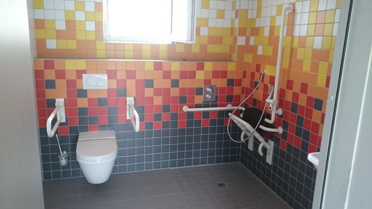 Ein modernes Badezimmer mit bunten Fliesen in Rot- und Gelbtönen. Es ist mit Haltegriffen und einem barrierefreien WC ausgestattet.