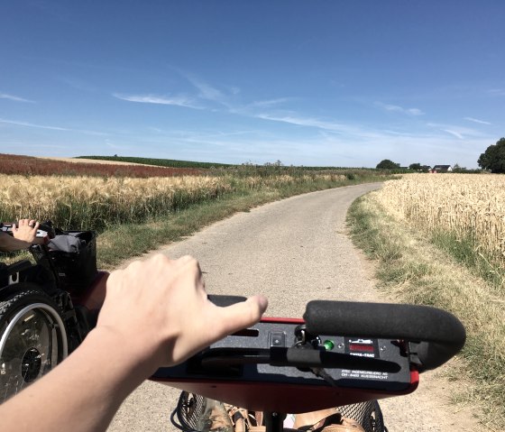 A person drives a Swiss Trac on a rural road, surrounded by golden and green fields under a clear blue sky., &copy; TI Bitburger Land