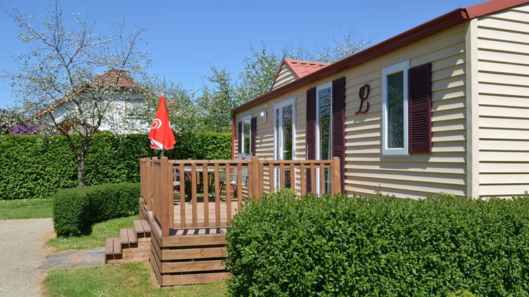 Ein gemütliches Mobilheim mit einer Holzterrasse und einem roten Fähnchen. Umgeben von grünen Hecken und einem klaren blauen Himmel.