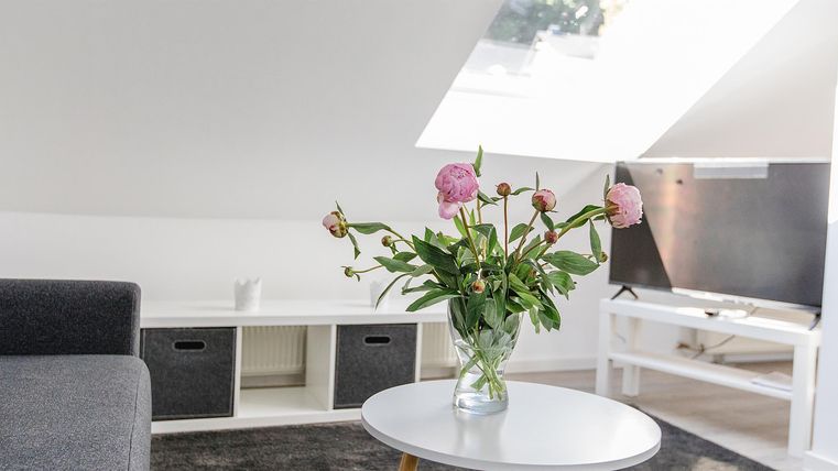 Ein heller Wohnraum mit einer modernen Couch und einem kleinen Tisch. Eine Vase mit bunten Blumen steht im Vordergrund, und ein Fernseher ist im Hintergrund zu sehen.