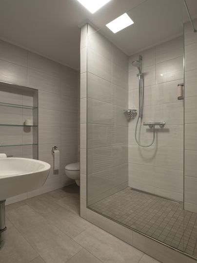 Ein modernes Badezimmer mit einer Duschkabine und einem Waschbecken. Die Wände sind in neutralen Tönen gefliest, wodurch der Raum hell und einladend wirkt.