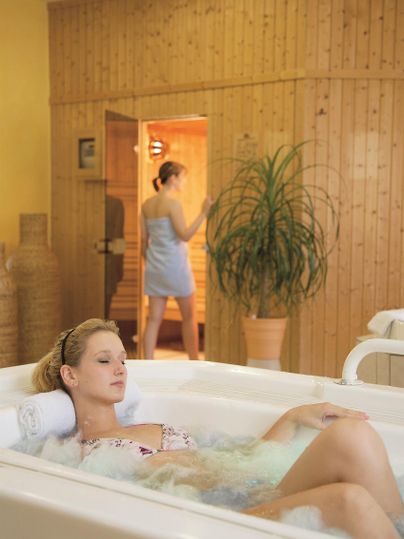 Een vrouw ontspant in een bubbelbad. Op de achtergrond betreedt een andere vrouw een sauna.