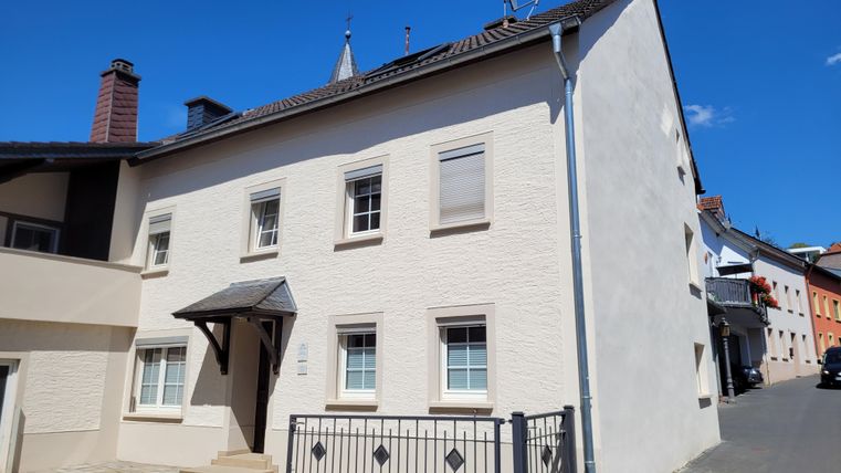 Ein modernes, helles Haus mit weißer Fassade. Neben dem Haus führt eine gepflasterte Straße mit angrenzenden Wohngebäuden.