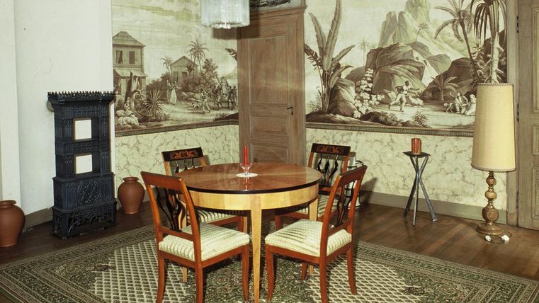 Ein stilvoll eingerichteter Raum mit einem runden Tisch und vier Stühlen. Die Wände sind mit Pflanzenmotiven dekoriert und eine stehlampe steht in der Ecke.