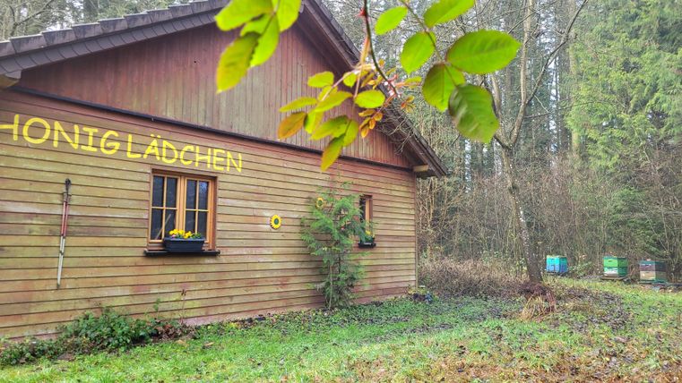 Een gezellig houten huis in een bosgebied met de tekst "Honiglädchen". Rondom het huis zijn weilanden en bomen te zien.