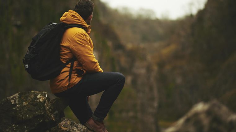 Ein Wanderer sitzt auf einem Felsen und schaut in eine tief eingeschnittene Schlucht. Er trägt eine gelbe Jacke und hat einen Rucksack dabei.