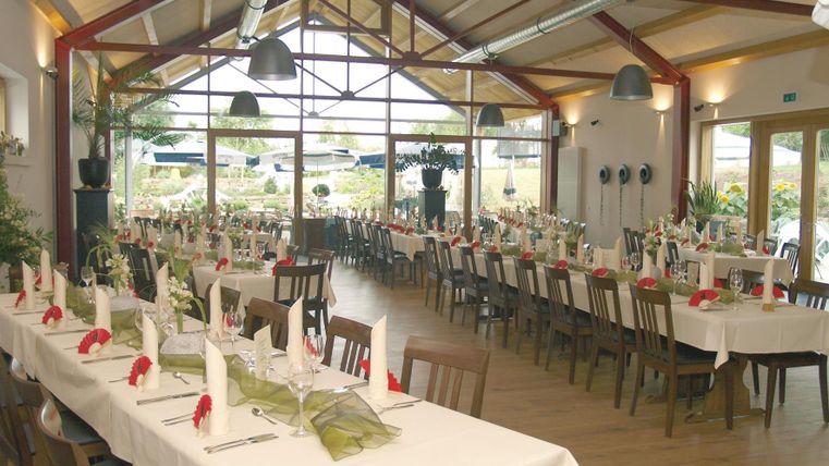 Ein heller und einladender Veranstaltungsraum mit langen Tischen, dekoriert mit Tischdecken und floralen Akzenten. Große Fenster bieten einen Blick auf die schöne Umgebung.