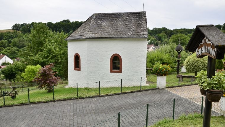 Eine kleine weiße Kapelle mit Schieferdach, umgeben von grüner Landschaft und einem Zaun, mit einem Schild "Kapellenhof" im Vordergrund.