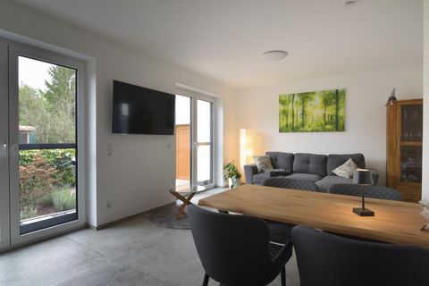 Een moderne woonkamer met een grijze bank en een houten tafel. Grote ramen laten veel licht binnen en een kunstwerk met groene bomen siert de muur.