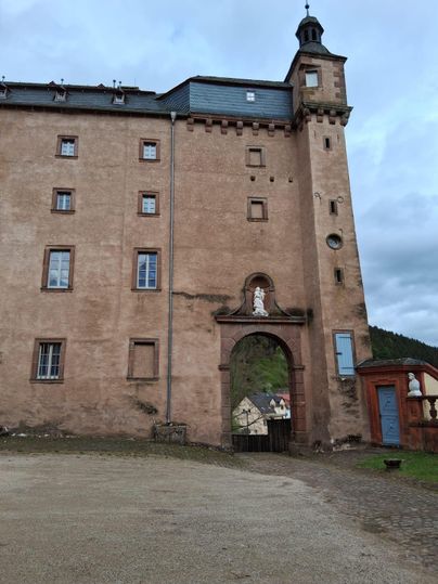 Un bâtiment historique avec des murs rustiques et une porte caractéristique. Au premier plan, des statues et une structure en pierre sont visibles.