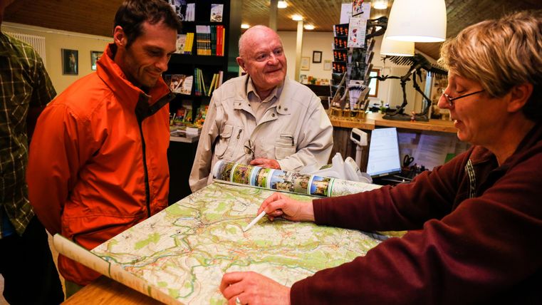 Een man in een oranje jasje en een oudere heer luisteren naar een vrouw die op een kaart wijst. De scène speelt zich af in een gezellige kamer die met verschillende voorwerpen is versierd.