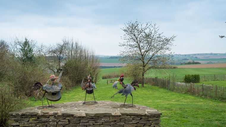 Een landelijke scène met meerdere sculpturen van vogels op een stenen plein. Op de achtergrond zijn weiden en een bewolkte lucht te zien.