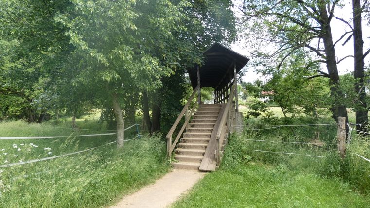 Überdachte Holzbrücke über einen kleinen Fluss, umgeben von Bäumen und Wiesen.