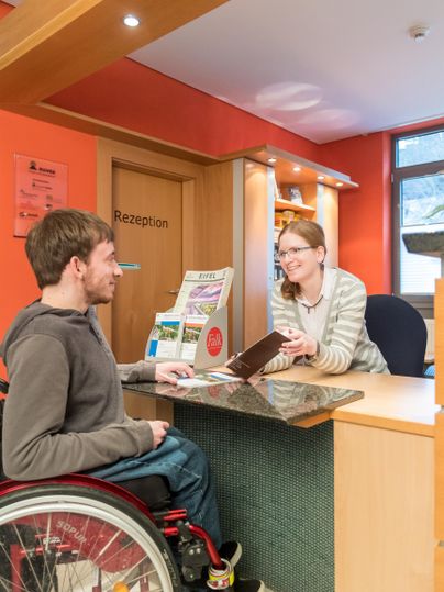 Een ontvangstgebied met een man in een rolstoel en een vrouw die aan de balie staat. Beiden lijken een vriendelijk gesprek te voeren.
