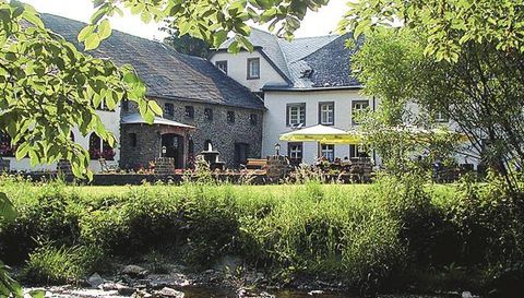 A charming house with a stone facade and large windows, surrounded by green meadows and trees. In the foreground, a small stream flows, and a terrace with sun umbrellas invites you to linger.