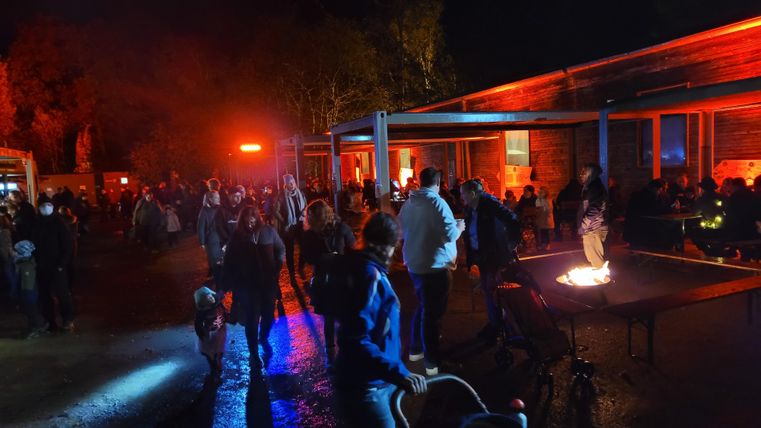 Eine lebhafte Nachtveranstaltung mit vielen Menschen, die sich unter bunten Lichtern versammeln. Im Vordergrund ist ein Lagerfeuer, das eine gemütliche Atmosphäre schafft.