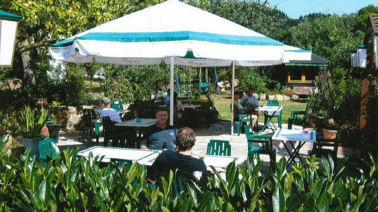 Ein gemütliches Café im Freien mit einem großen Sonnenschirm und grünen Stühlen. Umgeben von Pflanzen und Bäumen genießen die Gäste die entspannte Atmosphäre.